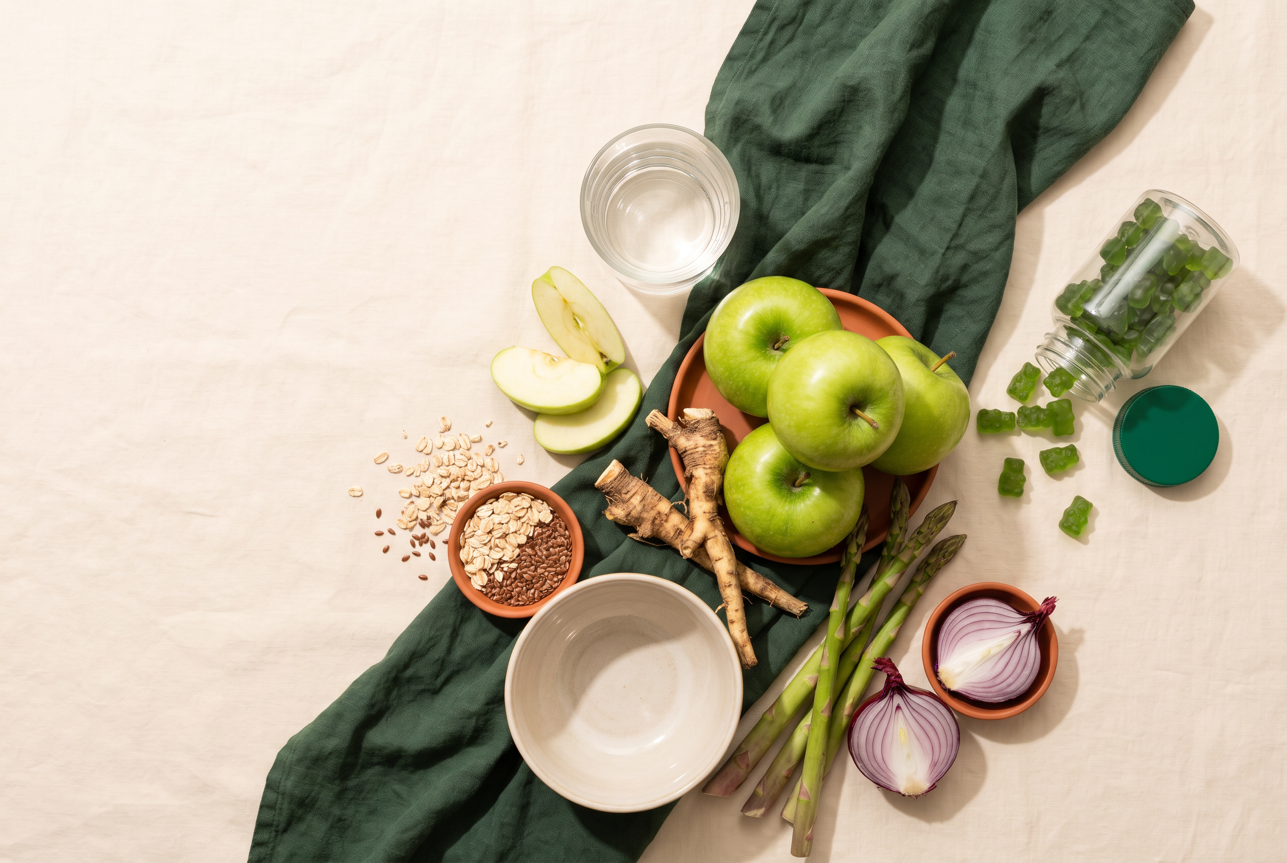 flat-lay של תפוחים ירוקים, שורש עולש, אספרגוס, שיבולת שועל ופשתן לצד כוס מים ובקבוק דובוני גומי בפלטת שמנת וירוק, בתאורת בוקר טבעית רכה